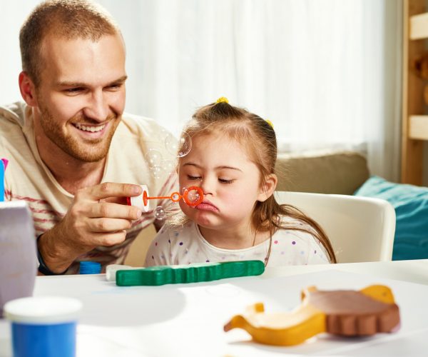 Happy young father holding bubble wand while his daughter with downs syndrome blowing soap bubbles at home